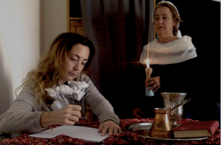 L'image montre deux femmes dans un environnement intime. L'une, assise, a les cheveux longs et bouclés, et elle semble concentrée sur l'écriture sur une feuille de papier. Elle porte un pull gris et une écharpe. L'autre femme se tient debout, légèrement en arrière, tenant une bougie allumée dans une main. Elle a les cheveux courts et une expression sérieuse. La pièce est faiblement éclairée, ce qui crée une ambiance chaleureuse. Sur la table, on peut voir un plateau avec une petite théière en métal et d'autres objets, ajoutant une touche rustique à la scène.