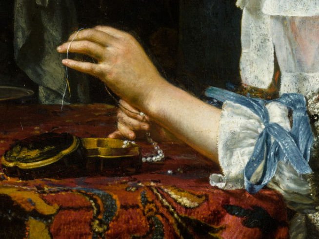 L'image montre une main délicate tenant un fil, probablement en train de travailler sur un collier de perles. Le poignet est orné d'un ruban bleu et la main semble concentrée sur l'activité. Une boîte ou un objet rond en bois est visible sur la table, ainsi qu'une étoffe ornée de motifs richement colorés, créant une atmosphère élégante.