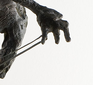 L'image semble représenter une partie d'une sculpture, mettant en avant une main réaliste et détaillée. La main est légèrement courbée et tendue, avec des doigts qui semblent interagir avec un fil ou une corde. La texture de la sculpture suggère un travail minutieux, probablement en métal ou en bronze, avec des ombres qui ajoutent de la profondeur. Le fond est clair, ce qui permet de mettre en valeur la forme et les détails de la main.