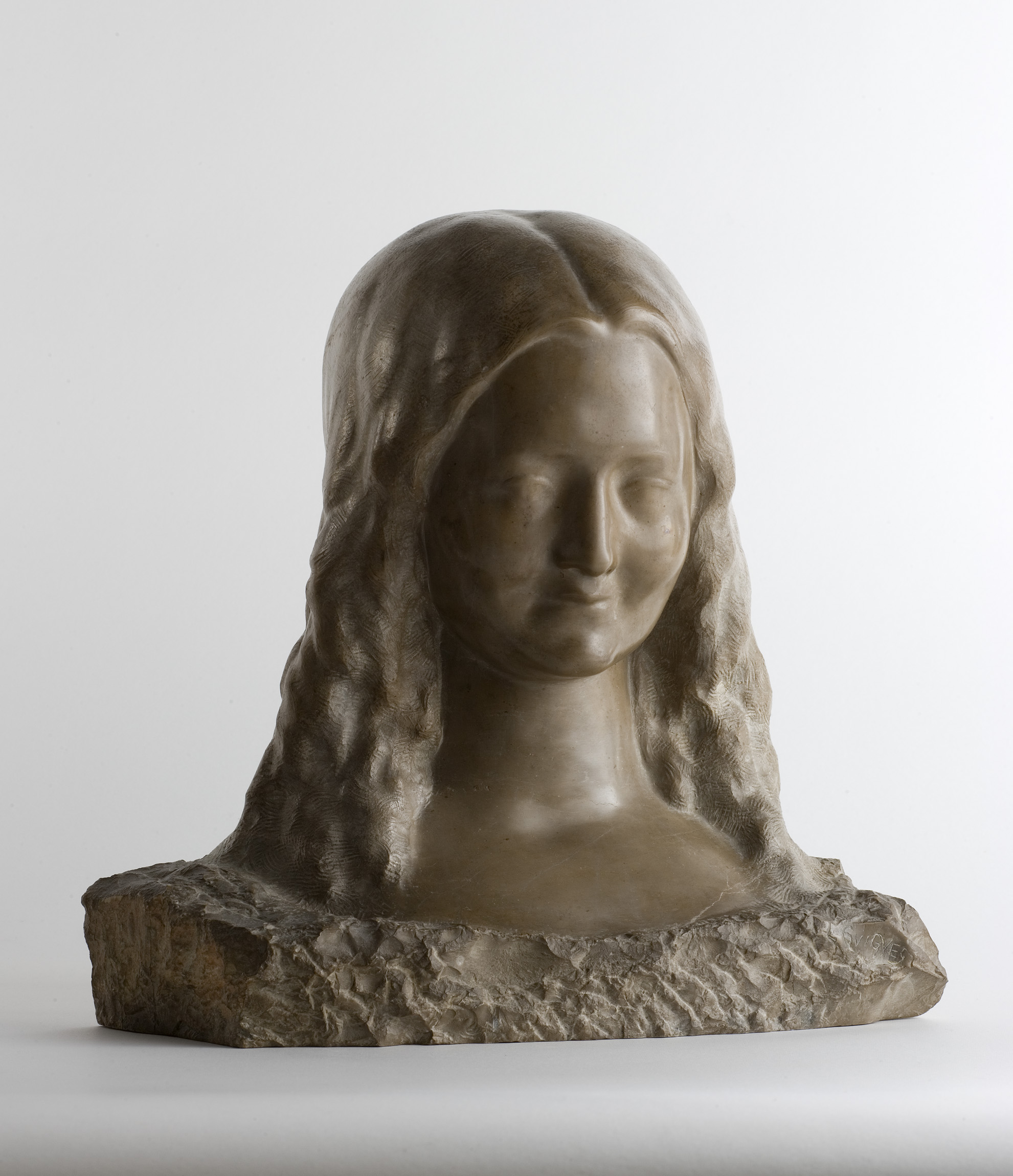 L'image montre une sculpture en pierre représentant un buste de femme. La sculpture met en avant le visage doux et serein de la femme, avec des cheveux ondulés qui tombent sur ses épaules. La texture de la pierre donne un aspect naturel et artistique, tandis que la base sur laquelle repose le buste est plus rugueuse, contrastant avec la douceur du visage. L'éclairage met en valeur les contours et les détails de la sculpture.