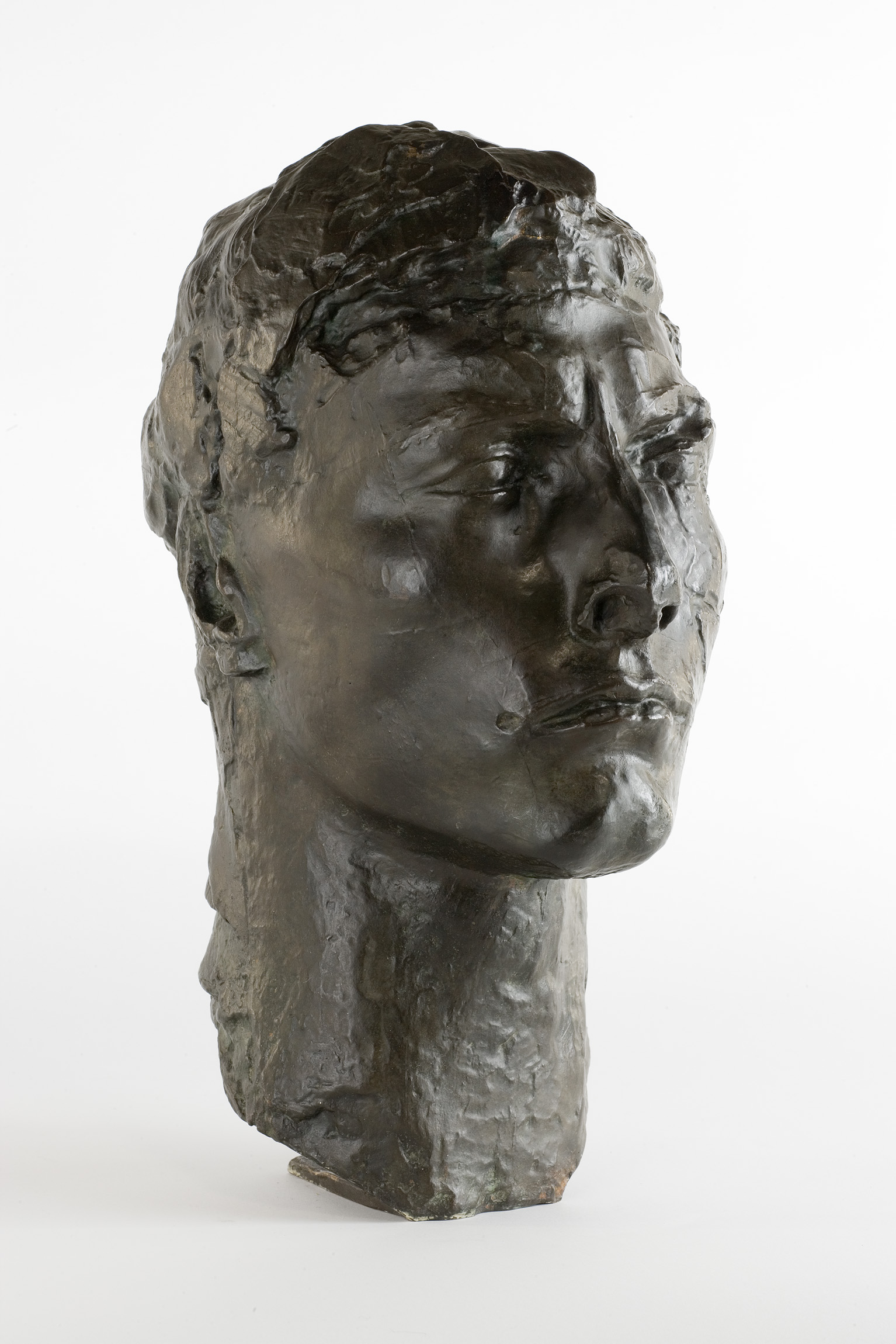 L'image montre une sculpture en bronze représentant un buste humain. Le visage est sculpté de manière réaliste, avec des traits prononcés et une expression intense. La surface de la sculpture présente une texture riche, typique du travail du bronze, et le fond est neutre, mettant en valeur la forme et les détails de l'œuvre. Cette pièce pourrait évoquer des thèmes liés à l'expression humaine ou à la condition humaine.