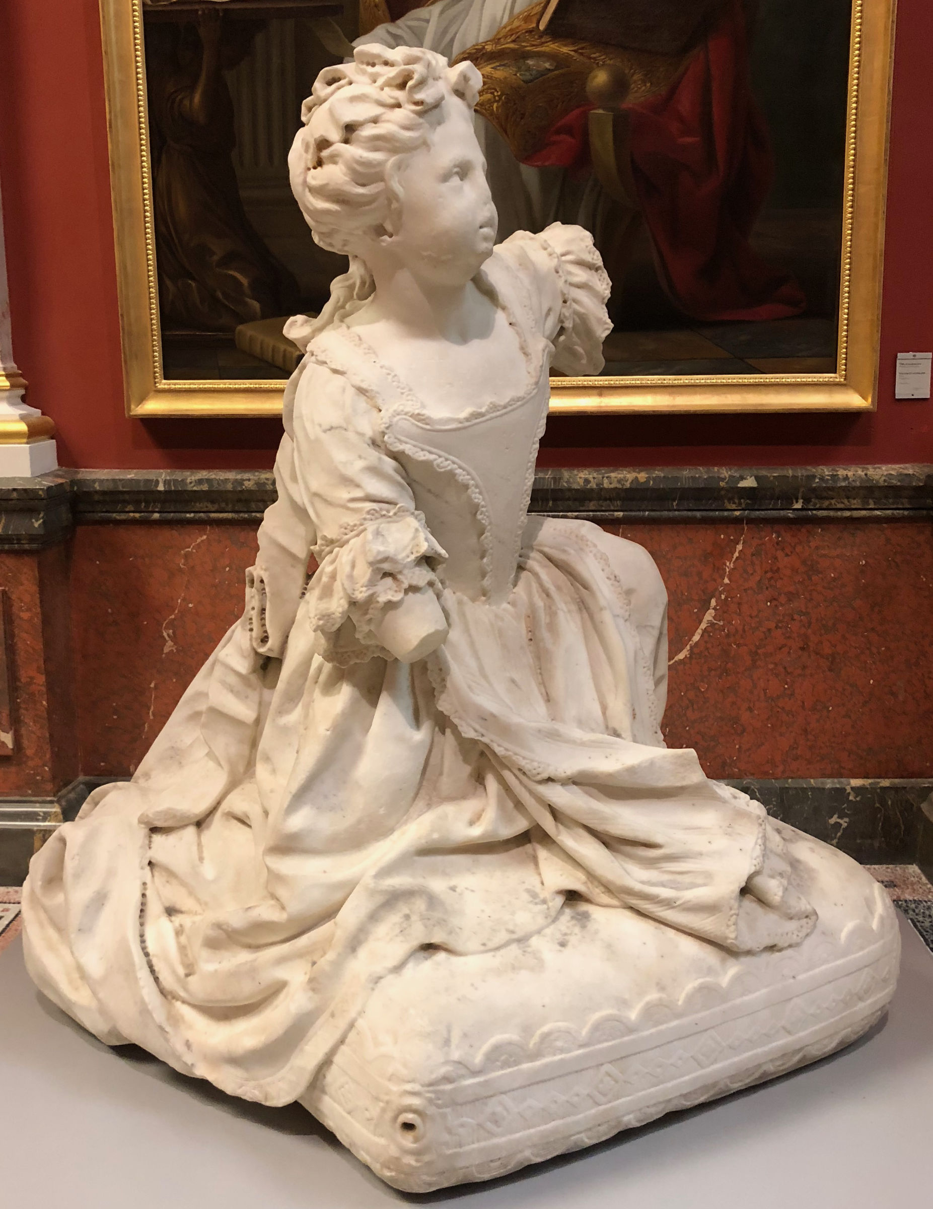 L'image montre une sculpture en marbre représentant une jeune fille assise. Elle est vêtue d'une robe élaborée avec des détails fins, et sa posture est élégante. Ses cheveux sont stylisés en boucles. La sculpture est placée sur une base moelleuse, ce qui accentue son apparence délicate. En arrière-plan, on peut voir une œuvre d'art, ce qui suggère que cette sculpture est exposée dans un musée.