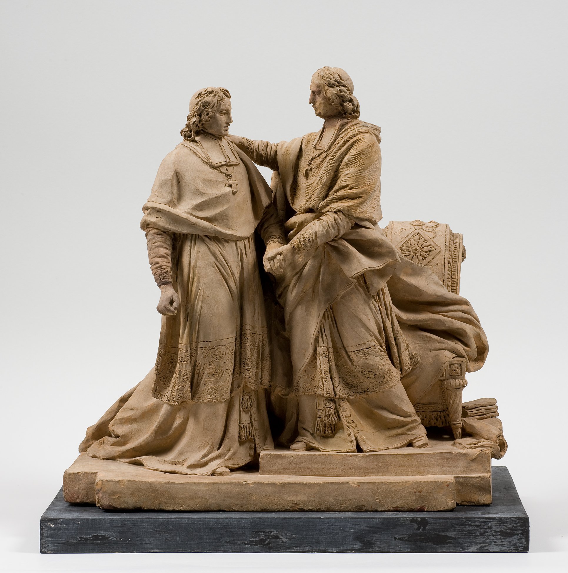 Cette image présente une sculpture en terre cuite représentant deux figures humaines en tenue historique. Les personnages sont debout, se faisant face, l'un touchant l'autre d'une manière affectueuse ou significative. Ils portent des vêtements richement détaillés, avec des drapés montrant un certain mouvement. Le socle est en pierre, ajoutant à la stabilité de l'œuvre. L'ensemble évoque une ambiance de proximité et d'intimité entre les deux figures.