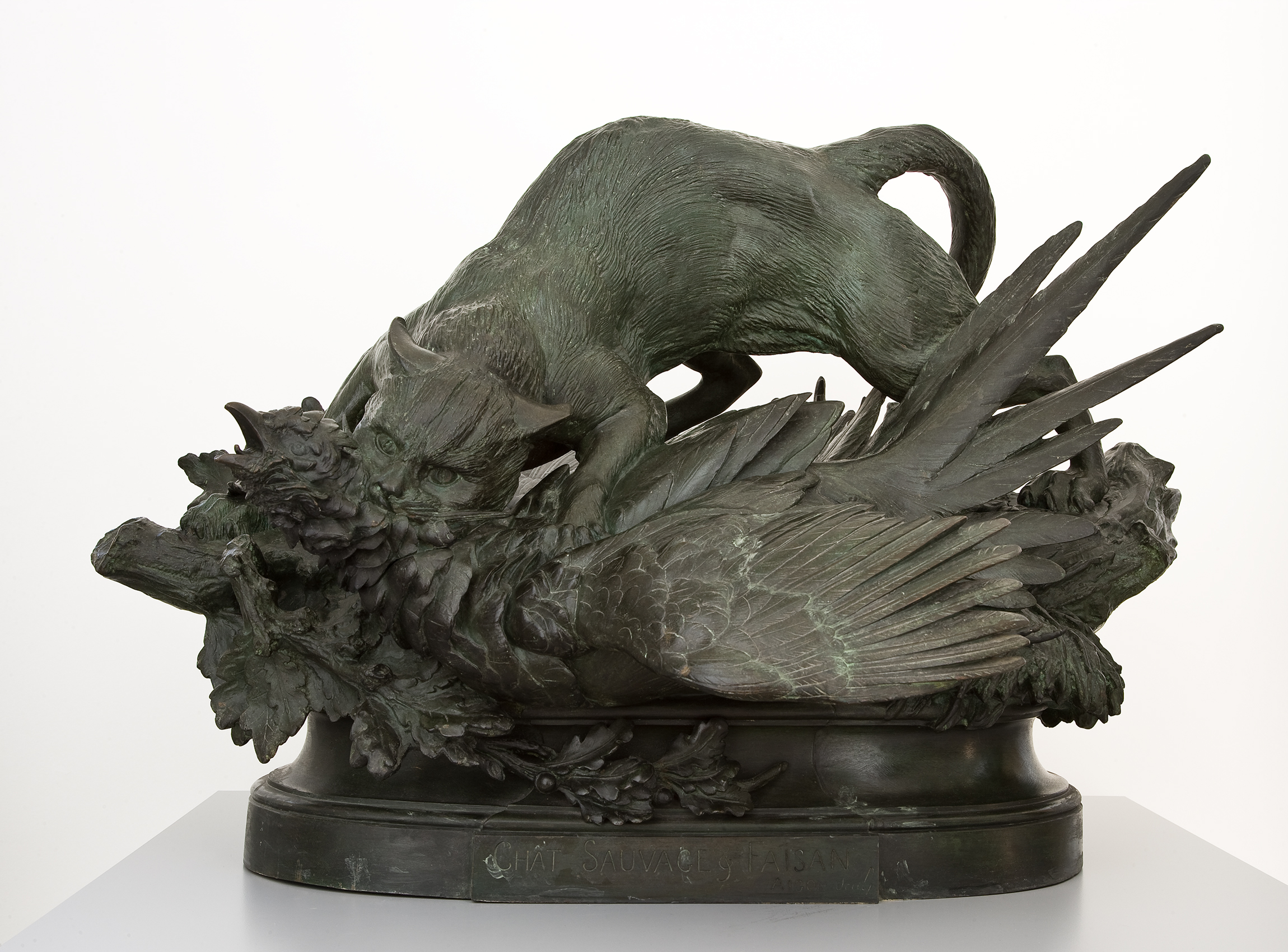 L'image représente une sculpture en bronze d'un chat en train de s'attaquer à un oiseau. Le chat, en position dynamique, semble être en mouvement, avec des détails précis sur le pelage. L'oiseau est également bien représenté, avec ses ailes déployées et des détails sur son plumage. La base de la sculpture est ornée de feuillages, ce qui ajoute une dimension naturelle à l'œuvre. L'ensemble dégage une atmosphère de chasse et de lutte, capturant un moment de la nature de manière artistique.