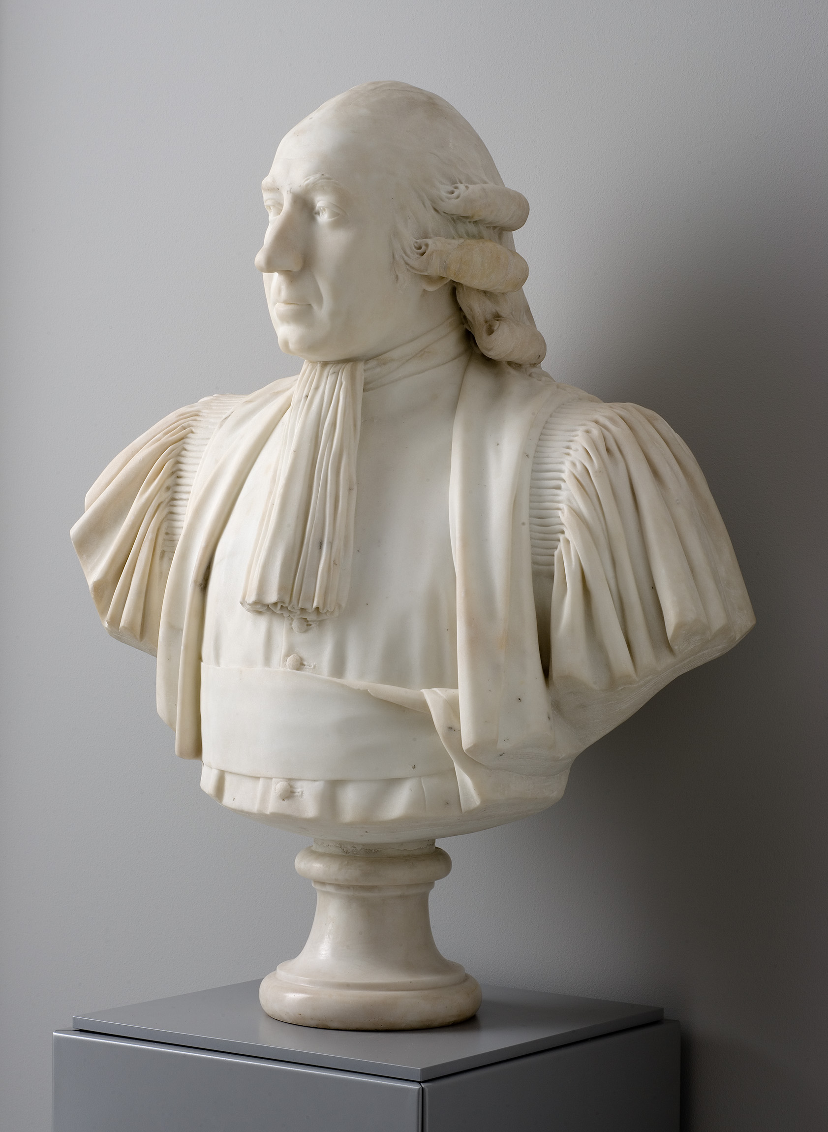 Cette image montre une sculpture en marbre représentant un buste d'homme. Le personnage est vêtu d'une chemise avec des manches plissées et porte une cravate. La sculpture est assez réaliste, mettant en valeur les traits du visage, les cheveux coiffés de manière élégante et le sérieux de l'expression. Le buste repose sur un socle, et l'ensemble est présenté dans un environnement minimaliste, ce qui permet de focaliser l'attention sur l'œuvre elle-même.