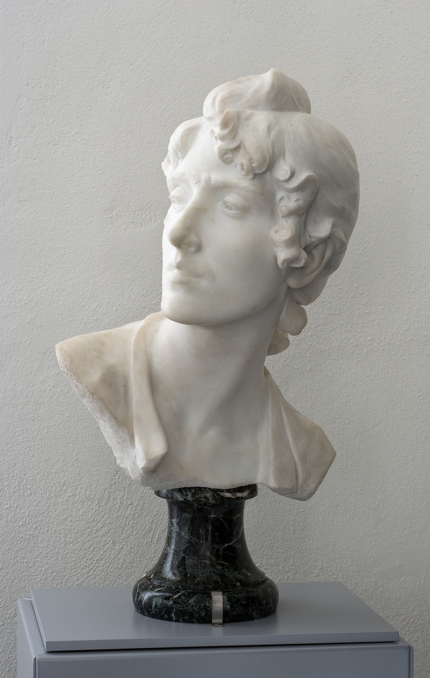 L'image montre une sculpture en marbre d'un buste masculin. Le personnage a une expression calme, avec des cheveux bouclés et une posture légèrement tournée. La sculpture est montée sur un piédestal en marbre noir, créant un bon contraste avec le marbre clair du buste. L'arrière-plan est sobre, mettant en valeur l'œuvre.