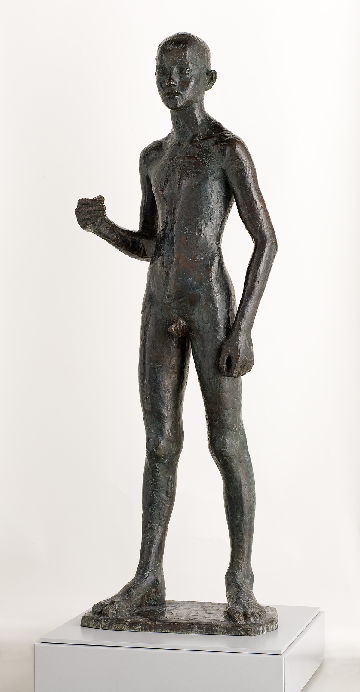 Cette image représente une sculpture en bronze d'une figure humaine debout. La statue est de taille réelle et montre un jeune homme nu, avec des détails réalistes dans la morphologie du corps. Le personnage se tient droit, légèrement incliné vers l'avant, avec un bras levé, tenant quelque chose dans sa main. Sa posture semble à la fois dynamique et sereine, et la texture de la surface métallique donne un aspect brut et organique à l'œuvre. Le socle sur lequel la sculpture repose est simple et met en valeur la figure.