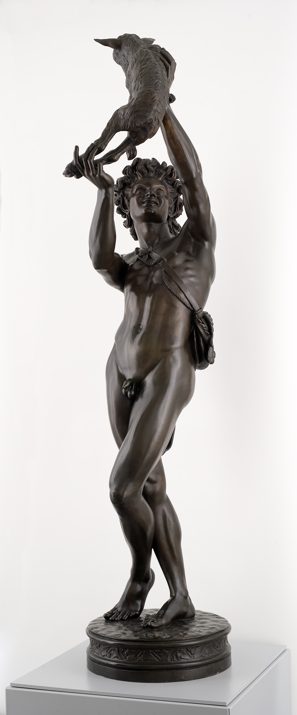L'image représente une sculpture en bronze d'un homme nu se tenant debout. Il porte une couronne de feuilles, symbolisant peut-être la fertilité ou la nature. Dans sa main droite, il soulève un oiseau, tandis que sa main gauche semble être posée sur son corps. La sculpture est réaliste et exprime une certaine dynamique dans la posture du personnage, qui semble à la fois puissant et en harmonie avec la nature. La base de la sculpture est ornée, ajoutant à son aspect artistique.