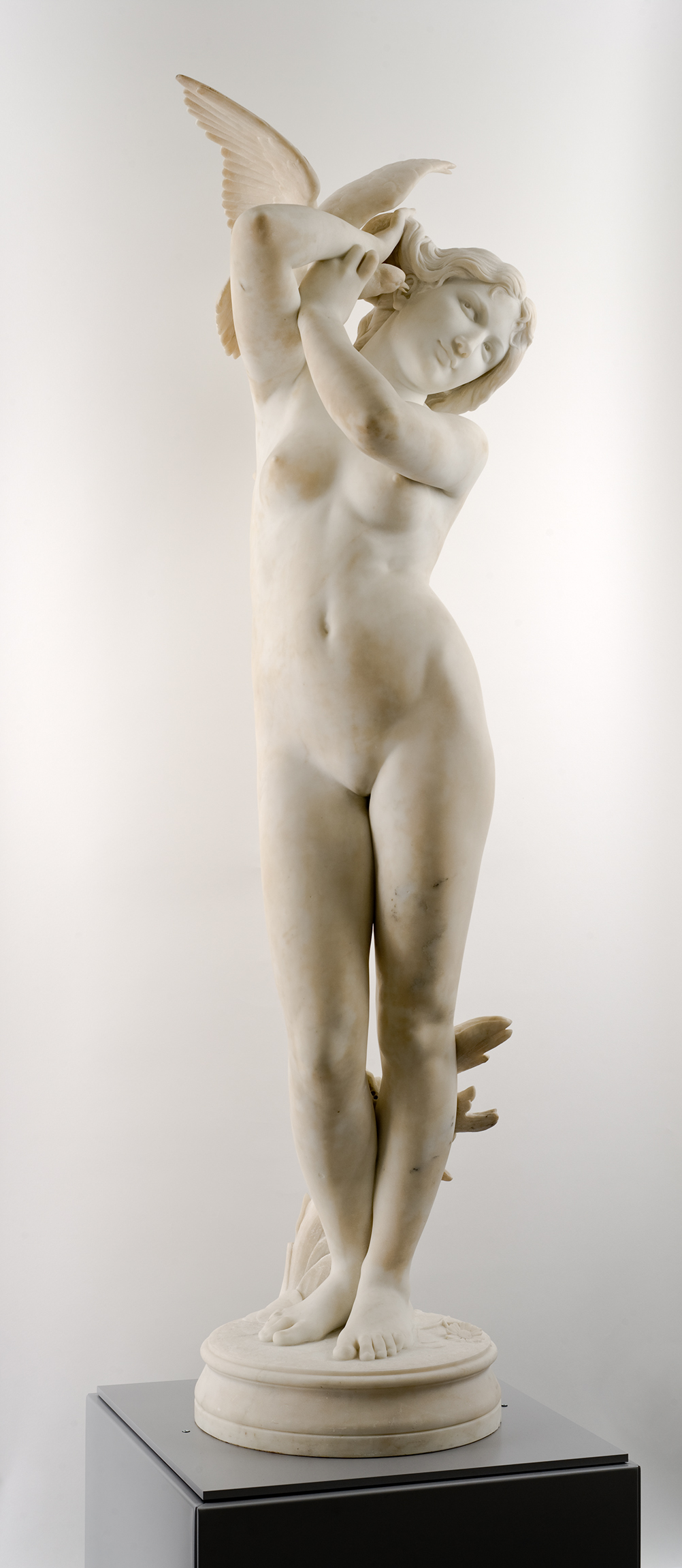 Cette image montre une sculpture en marbre d'une femme nue, représentant une figure féminine gracieuse. Elle se tient debout, légèrement tournée, avec une posture élégante. Ses bras sont levés, tenant délicatement une représentation d'un oiseau. La sculpture met en valeur des détails anatomiques soignés et une expression douce. Au bas de l'œuvre, il y a des éléments décoratifs qui semblent faire référence à la nature. Dans l'ensemble, l'œuvre dégage une impression de légèreté et de beauté.