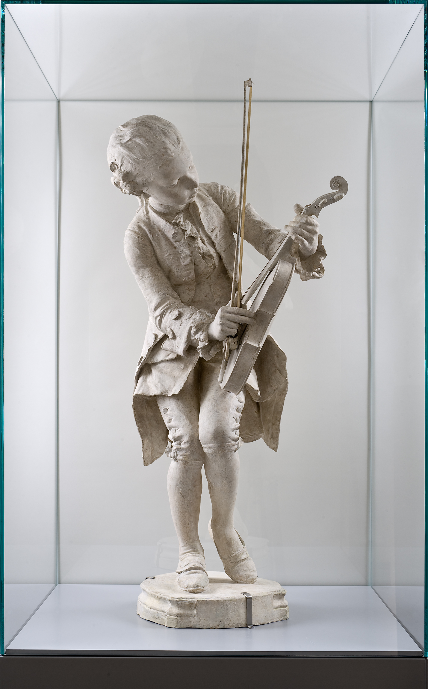 L'image montre une sculpture en marbre d'un jeune musicien jouant d'un instrument à cordes. Le personnage est représenté en train de jouer, avec une posture dynamique et un regard concentré. Il porte des vêtements d'époque, typiques du XVIIIe siècle, avec un costume richement détaillé. La sculpture est exposée dans une vitrine, mettant en avant la finesse des détails et le savoir-faire de l'artiste. Les ombres et les lumières soulignent les traits du visage et les plis des vêtements, ajoutant du réalisme à l'œuvre.