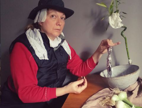L'image montre une femme assise à une table. Elle porte un chapeau noir et une écharpe en tissu blanc sur la tête. Elle a l'air concentrée et tient un objet en aluminium dans une main, tandis que l'autre main est posée sur la table. À côté d'elle, il y a un bol en pierre et une plante de bambou décorative, ainsi qu'un tissu sur la table, avec quelques légumes, peut-être des oignons. L'éclairage est doux, donnant une ambiance calme à l'ensemble.