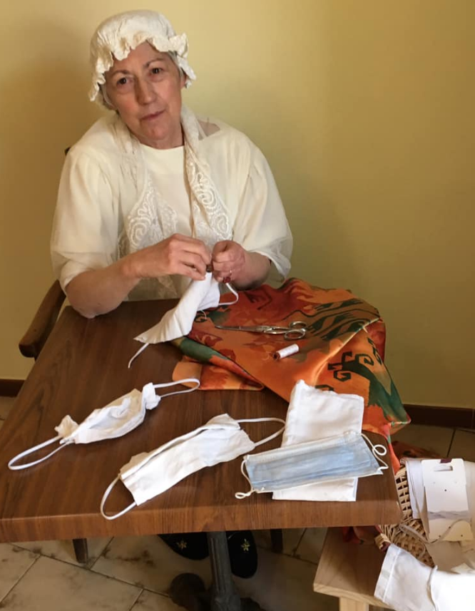 L'image montre une femme assise à une table, portant une coiffe et un vêtement traditionnel. Elle semble en train de coudre ou de réparer des masques en tissu. Sur la table, on peut voir plusieurs masques et des outils de couture, ainsi qu'un tissu coloré. L'arrière-plan est d'une couleur neutre, ce qui met en valeur l'activité qu'elle réalise.