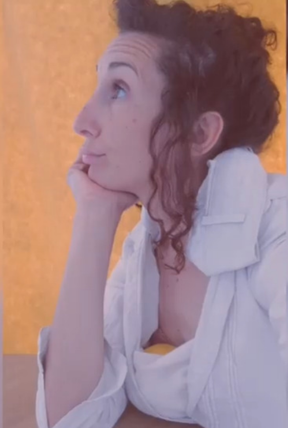 L'image montre une femme avec des cheveux bouclés, posant de profil. Elle a l'air pensif, avec une main soutenant son menton. Elle porte une chemise blanche, et l'arrière-plan est de couleur jaune vif, ce qui crée un contraste intéressant. Son expression faciale semble douce et contemplative.