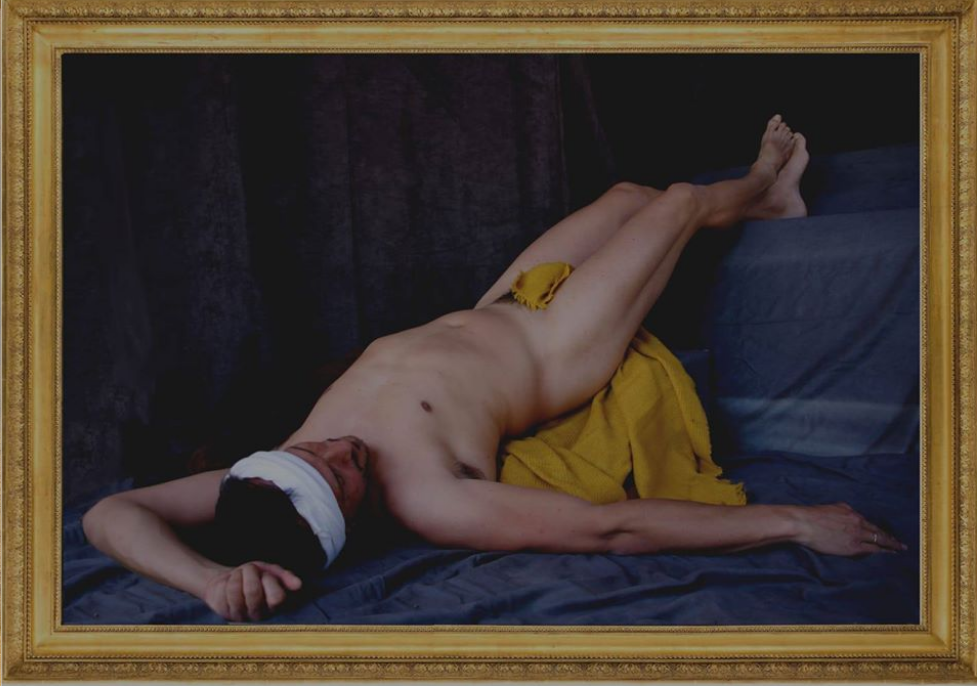 L'image représente un homme allongé sur une surface grise. Il est nu, avec un bandeau blanc autour de la tête et un petit morceau de tissu jaune couvrant partiellement son corps. Son bras est étendu, et il semble être dans une pose relaxée, avec une expression neutre. L'arrière-plan est sombre, ce qui met en évidence la chair et les tissus colorés. L'ensemble a une composition artistique et contemplative.