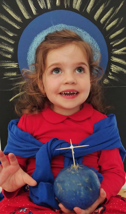 L'image montre une petite fille avec de longs cheveux bouclés, vêtue d'une chemise rouge et d'une écharpe bleue. Elle tient une pomme ou un fruit bleu dans ses mains, avec une petite brochette plantée dessus. Elle a un sourire joyeux et semble émerveillée. En arrière-plan, il y a un dessin représentant un cercle bleu et des rayons, créant une ambiance lumineuse. La scène semble festive et ludique.