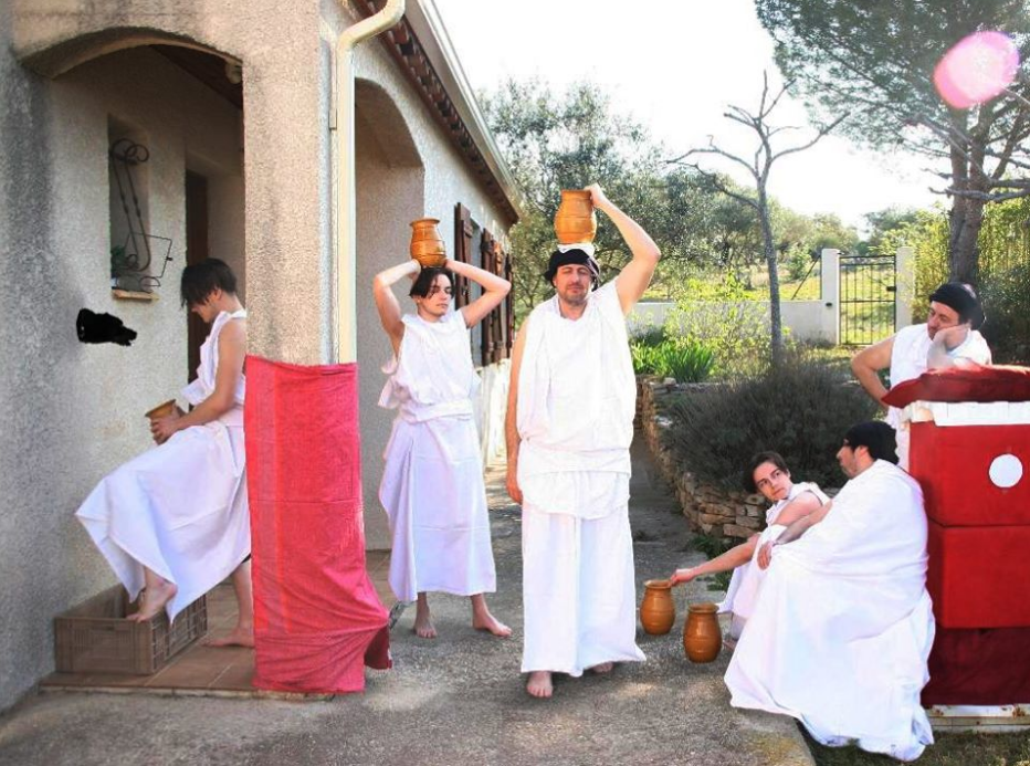L'image montre un groupe de personnes, vêtues de toges blanches, qui semblent se préparer pour une scène ou un événement inspiré de l'Antiquité. Certains d'entre eux portent des pots en terre sur la tête, tandis que d'autres sont assis ou se tiennent debout près d'un mur d'une maison. L'environnement est extérieur, avec des arbres en arrière-plan et une ambiance ensoleillée. L'ensemble donne une impression de convivialité et de mise en scène historique.