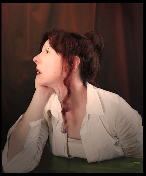 L'image montre une femme dans une pose contemplative. Elle a les cheveux bruns relevés en un chignon et porte une chemise blanche. Son visage est légèrement tourné sur le côté, ce qui donne une impression de réflexion ou de rêve. Le fond est sombre, mettant en valeur la silhouette et l'expression de la femme. L'éclairage semble doux, créant une atmosphère intime.