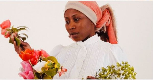 L'image montre une femme portant un haut blanc et un foulard. Elle tient des bouquets de fleurs colorées dans ses mains et semble contemplative, portant son regard sur les fleurs. Le fond de l'image est clair, mettant en valeur sa tenue et les fleurs qu'elle présente. L'ambiance générale transmet une sensibilité à la nature et à la beauté des fleurs.