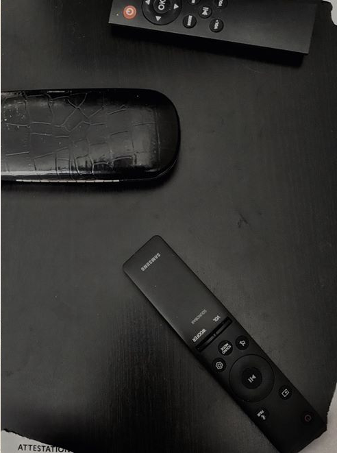 L'image montre une surface noire sur laquelle se trouvent plusieurs objets. On voit une télécommande de marque Samsung, ainsi qu'une autre télécommande ou un dispositif à côté d'elle. Il y a aussi des lunettes noires, posées sur la même surface. L'ensemble de l'image présente une ambiance sobre, avec un fond minimaliste.