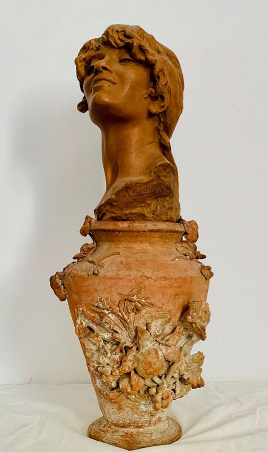 L'image montre une sculpture en terre cuite représentant un buste féminin. La tête de la femme est élégamment posée sur un grand vase orné de motifs floraux détaillés. Le vase est de couleur terre cuite, avec des nuances de brun et d'orange, tandis que le buste présente des traits fins et une expression contemplative. Les fleurs sculptées sur le vase ajoutent une touche artistique à l'ensemble, créant une harmonie entre le buste et le support. L'ensemble dégage une impression de délicatesse et d'élégance.
