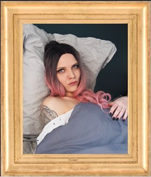 L'image montre une femme allongée sur un lit avec des draps grisés. Elle a les cheveux longs, teintés de rose, et fixe l'objectif avec un regard intense. Son bras est légèrement visible, montrant un tatouage. Le cadre en bois doré accentue l'aspect artistique de la photographie. L'ambiance générale est à la fois intime et réfléchie.