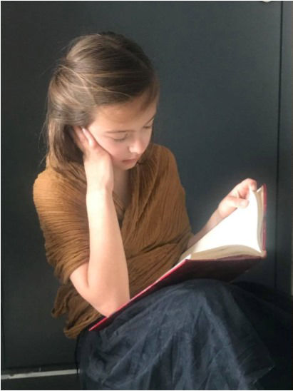 L'image montre une jeune fille assise, concentrée sur un livre. Elle a les cheveux châtain clair et porte un vêtement marron. Sa posture indique qu'elle est absorbée par sa lecture, tenant le livre d'une main et posant l'autre main sur son visage. L'arrière-plan est sombre, ce qui fait ressortir son expression et l'objet de son attention.
