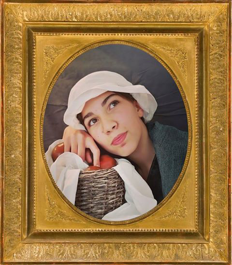 L'image représente une jeune personne, visiblement dans un style historique, portant un chapeau blanc et une tenue traditionnelle. Elle est assise près d'un panier en osier rempli de fruits, probablement des pommes. Son expression semble rêveuse, les yeux tournés vers le haut, ce qui ajoute une touche de mélancolie à l'ensemble. Le tout est encadré dans un cadre doré, ce qui donne une impression de classicisme à la scène.