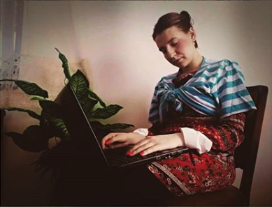 L'image montre une femme assise sur une chaise, concentrée sur un ordinateur portable. Elle porte une tenue colorée, avec un haut rayé et une robe à motifs floraux. À côté d'elle, on peut voir une plante verte qui ajoute une touche de nature à l'environnement. La lumière semble douce, créant une atmosphère calme et intime.