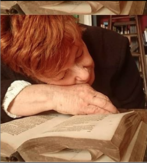 L'image montre une personne au visage légèrement penché, reposant sa tête sur un livre ouvert. Ses cheveux sont de couleur roux et elle porte un haut noir. L'atmosphère de la scène semble calme et contemplative, comme si la personne était plongée dans la lecture ou l'étude. Les pages du livre sont légèrement jaunies, ce qui donne une impression de vintage ou d'ancienneté à l'objet. L'arrière-plan suggère un environnement intérieur, peut-être une bibliothèque ou un espace de travail.