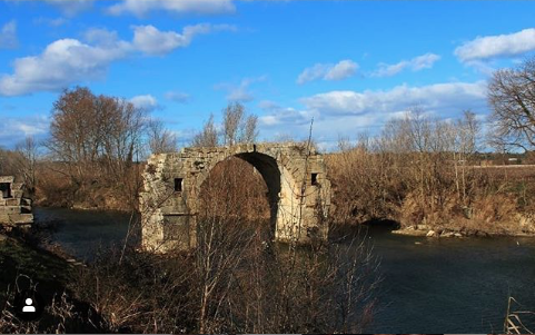 L'image montre un ancien pont en ruines, avec un arc majestueux au-dessus d'une rivière. La structure en pierre est partiellement dégradée, ce qui lui donne un aspect pittoresque. Autour du pont, on aperçoit des arbres sans feuillage, typiques de l'hiver, et un ciel bleu parsemé de nuages blancs. La scène évoque un sentiment de calme et de tranquillité, tout en mettant en valeur la beauté naturelle des environs.