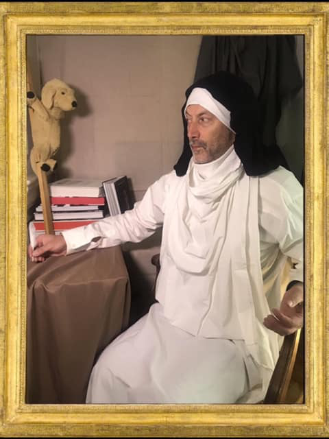 L'image montre un homme assis, vêtu d'une tenue blanche traditionnelle. Il porte un turban ou un foulard noir sur la tête et semble contemplatif. À côté de lui, un petit chien en peluche est posé sur une étagère avec des livres. Le décor a une ambiance sobre et l'homme est capturé dans une pose réfléchie, ce qui ajoute une touche intéressante à la scène. Le tout est encadré de manière artistique, ce qui donne un aspect théâtral à l'image.