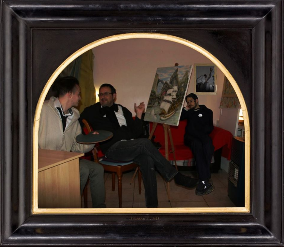 L'image montre un intérieur où trois hommes sont assis autour d'une table. L'un d'eux semble tenir une assiette, tandis qu'un autre est en train de discuter, peut-être en pointant quelque chose sur la toile qui est placée derrière eux. On peut également voir une peinture accrochée au mur, ainsi qu'un canapé rouge dans la pièce. L'ambiance semble conviviale et détendue. Le cadre de la photo est en bois noir, donnant un aspect artistique à l'image.