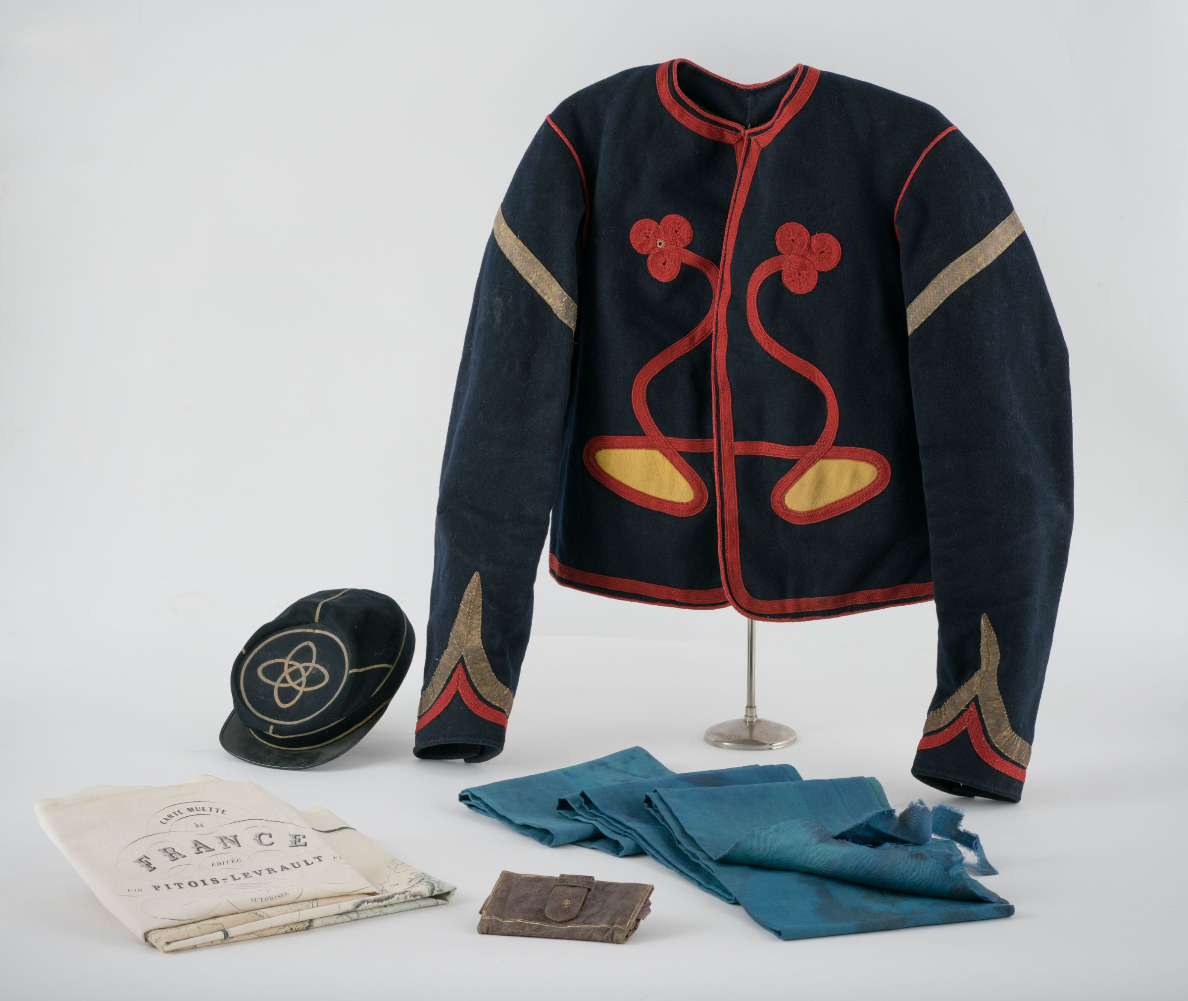 L'image montre une tenue historique, probablement militaire, avec une cape noire décorée de motifs rouges et dorés. À côté d'elle se trouvent divers objets, dont un chapeau noir avec un symbole, un document plié avec le mot "France" visible, ainsi qu'un petit portefeuille et des morceaux de tissu bleu. L'ensemble présente un aspect nostalgique et témoigne d'une période passée.