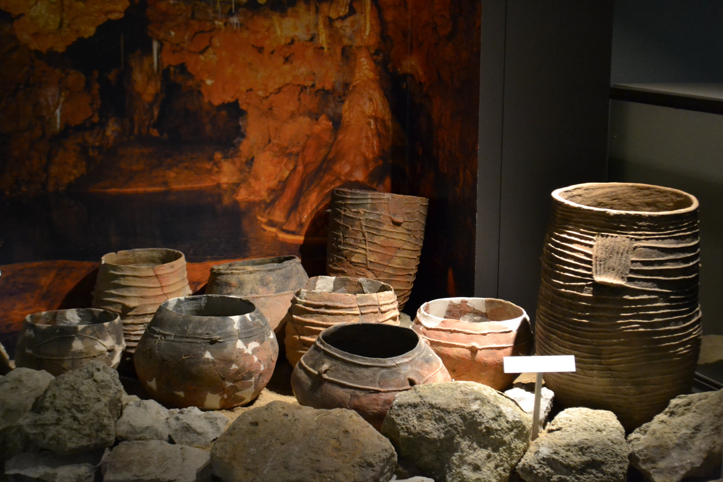 L'image montre une collection de poteries anciennes disposées sur une surface, entourées de pierres. Les pots ont différentes formes et textures, certains sont décorés avec des motifs. L'arrière-plan présente une teinte sombre qui évoque une ambiance archéologique, suggérant que ces objets sont exposés dans un musée ou un site historique. Les poteries illustrent la créativité et l'artisanat des civilisations passées.