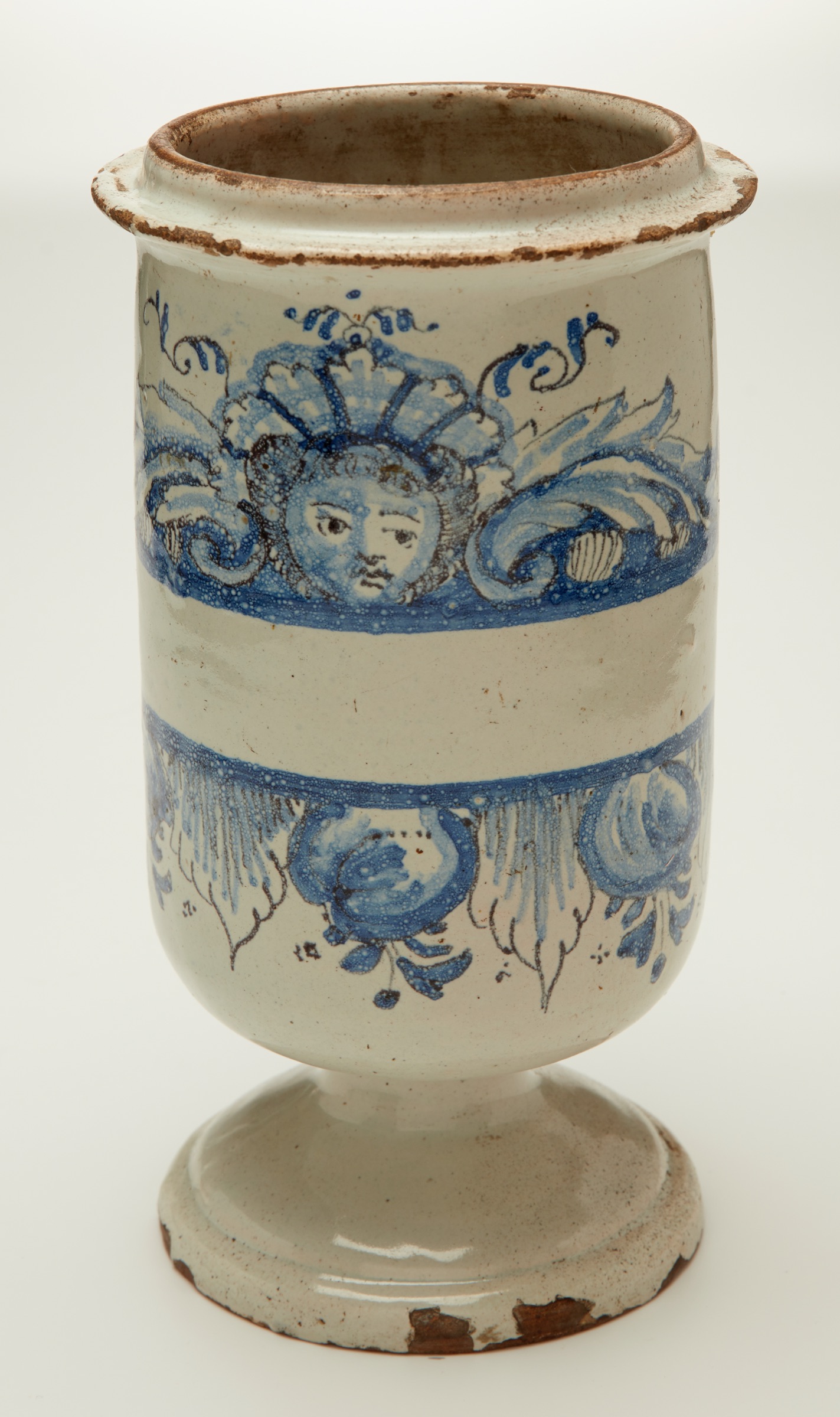 L'image montre un vase en céramique aux couleurs claires, notamment beige et bleu. Il est de forme élancée avec un socle ample et un rebord légèrement évasé. Sur la surface du vase, on peut observer des motifs décoratifs, dont un visage en bleu entouré de motifs floraux. La finition présente quelques imperfections, telles que des éraflures et des traces d'usure, ce qui lui confère un caractère antique et artisanal.