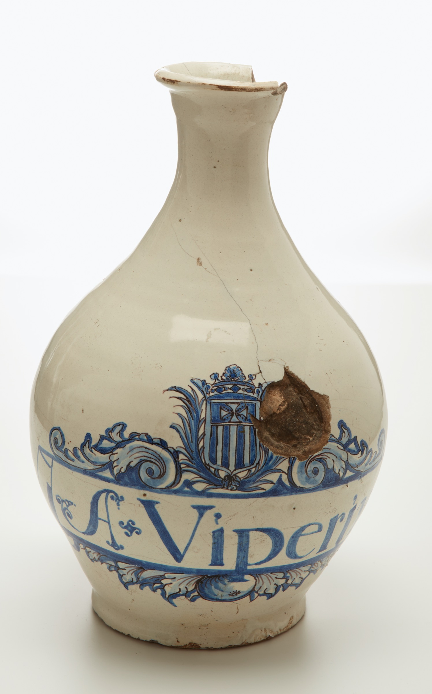 L'image montre une jarre en céramique de couleur blanche avec des motifs bleus. Elle présente une forme arrondie et étroite au niveau du goulot. Sur la surface, on peut voir des inscriptions et un blason, ainsi qu'un endroit où la jarre est endommagée, avec un trou visible. Les motifs décoratifs sont détaillés, ajoutant un aspect artistique à l'objet.