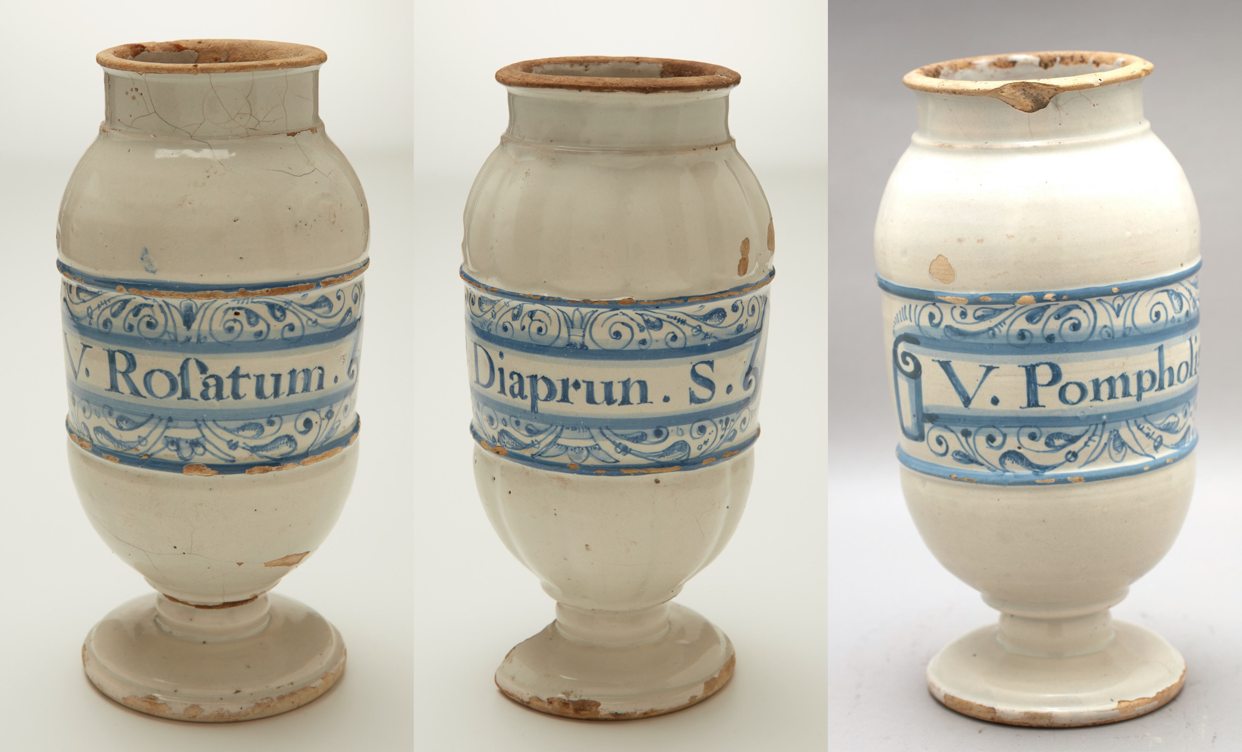 L'image présente trois angles d'une jarre ou vase en céramique. Ce récipient est de couleur blanche avec des décorations en bleu. Sur le bandeau bleu, on peut lire des inscriptions qui semblent nommer des substances ou remèdes, probablement d'origine médicale, comme "Rofatum", "Diaprun" et "Pomphol". La surface présente des imperfections et des éclats, témoignant de son âge et de son utilisation. La forme est stylisée avec un corps élancé et une base légèrement évasée.