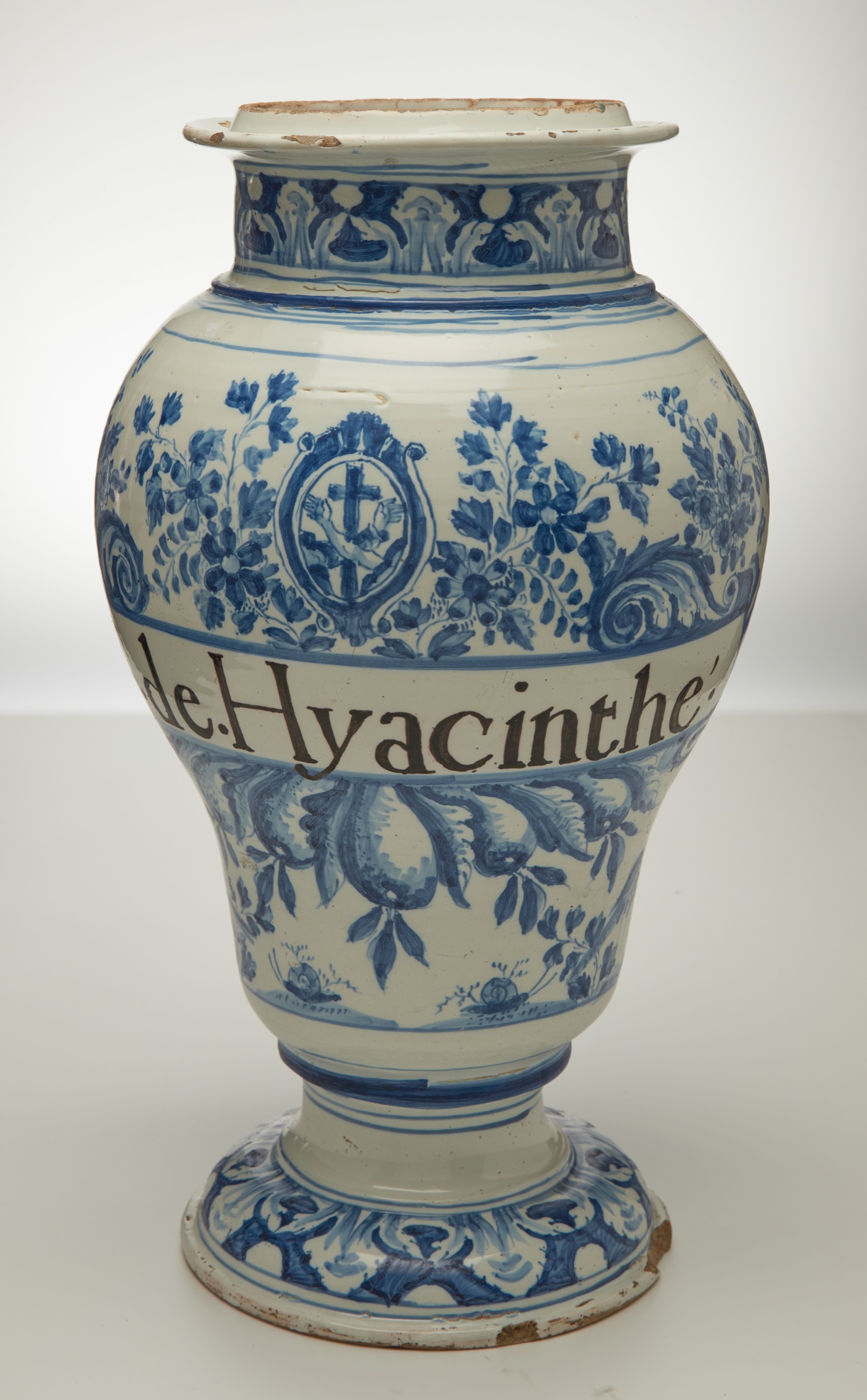 L'image montre un vase en céramique, décoré de motifs floraux en bleu sur fond blanc. Le vase a une forme classique, avec un col étroit et un corps arrondi, reposant sur une base plus large. Sur le vase, on peut voir une inscription en lettres foncées qui dénote probablement un nom ou une description. Les détails floraux et les motifs ornés ajoutent une touche d'élégance à l'objet.