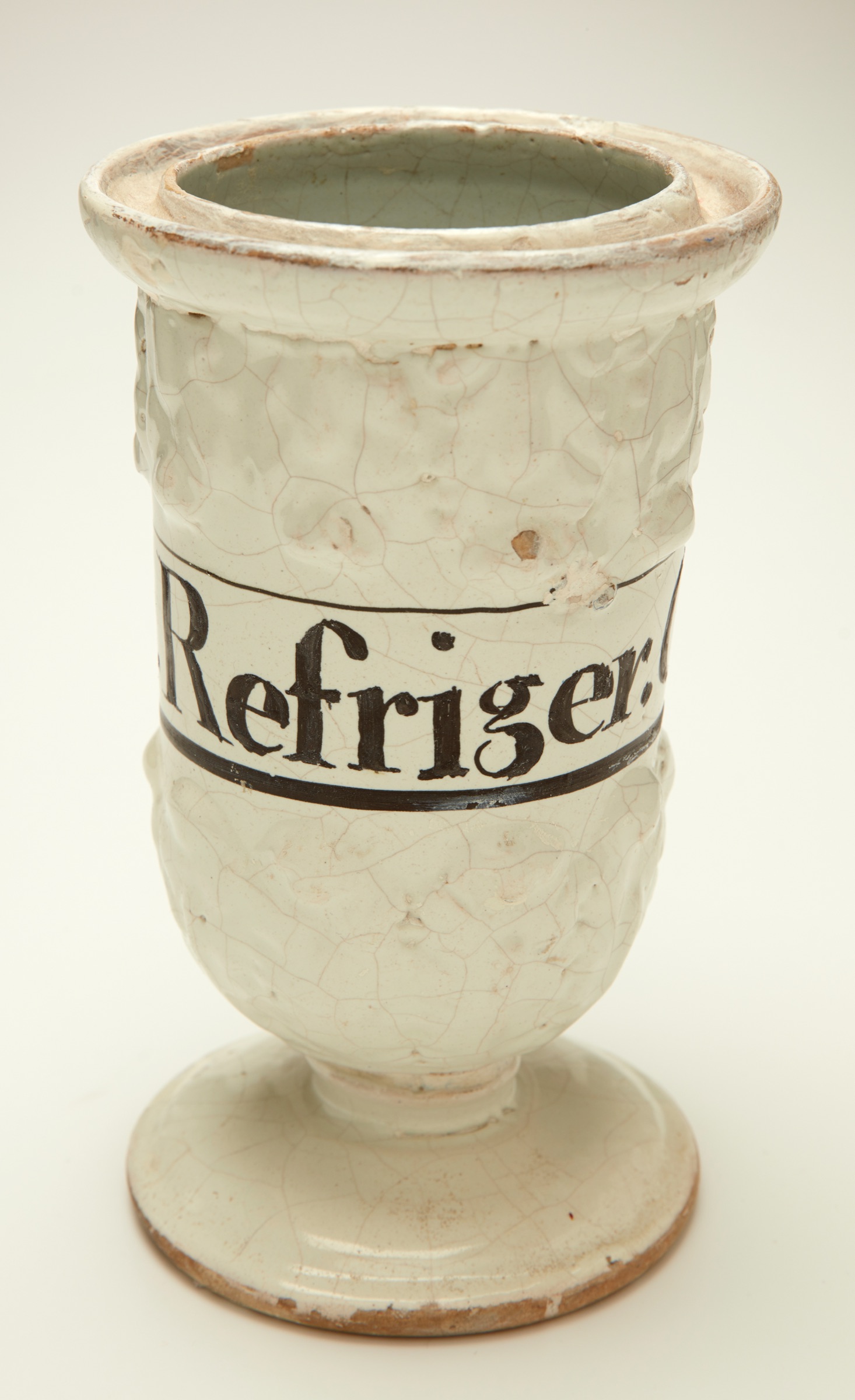 L'image représente un récipient en céramique décoratif, de forme cylindrique avec un pied. La surface est texturée et présente de légères craquelures, lui donnant un aspect ancien. Un bandeau sur le récipient porte le mot "Refriger," suggérant qu'il s'agit d'un objet destiné à conserver quelque chose au frais. L'ensemble dégage un charme vintage et artisanal.