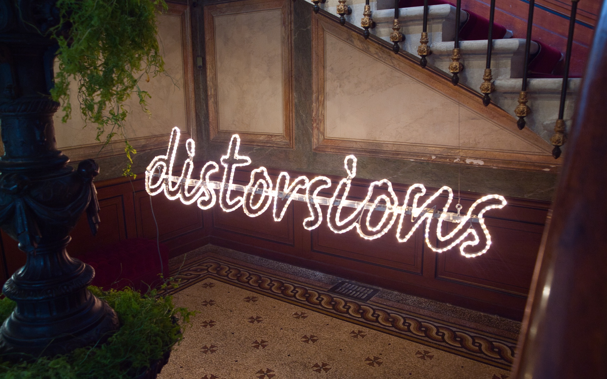 L'image présente un intérieur mettant en valeur un escalier. Sur le mur, des lumières en néon forment le mot "distortions" dans un style manuscrit. On peut également apercevoir des éléments décoratifs tels qu'une plante verte et un balustre en fer forgé, créant une ambiance à la fois élégante et artistique. Les matériaux comme le bois et la pierre ajoutent une touche classique à l'espace.