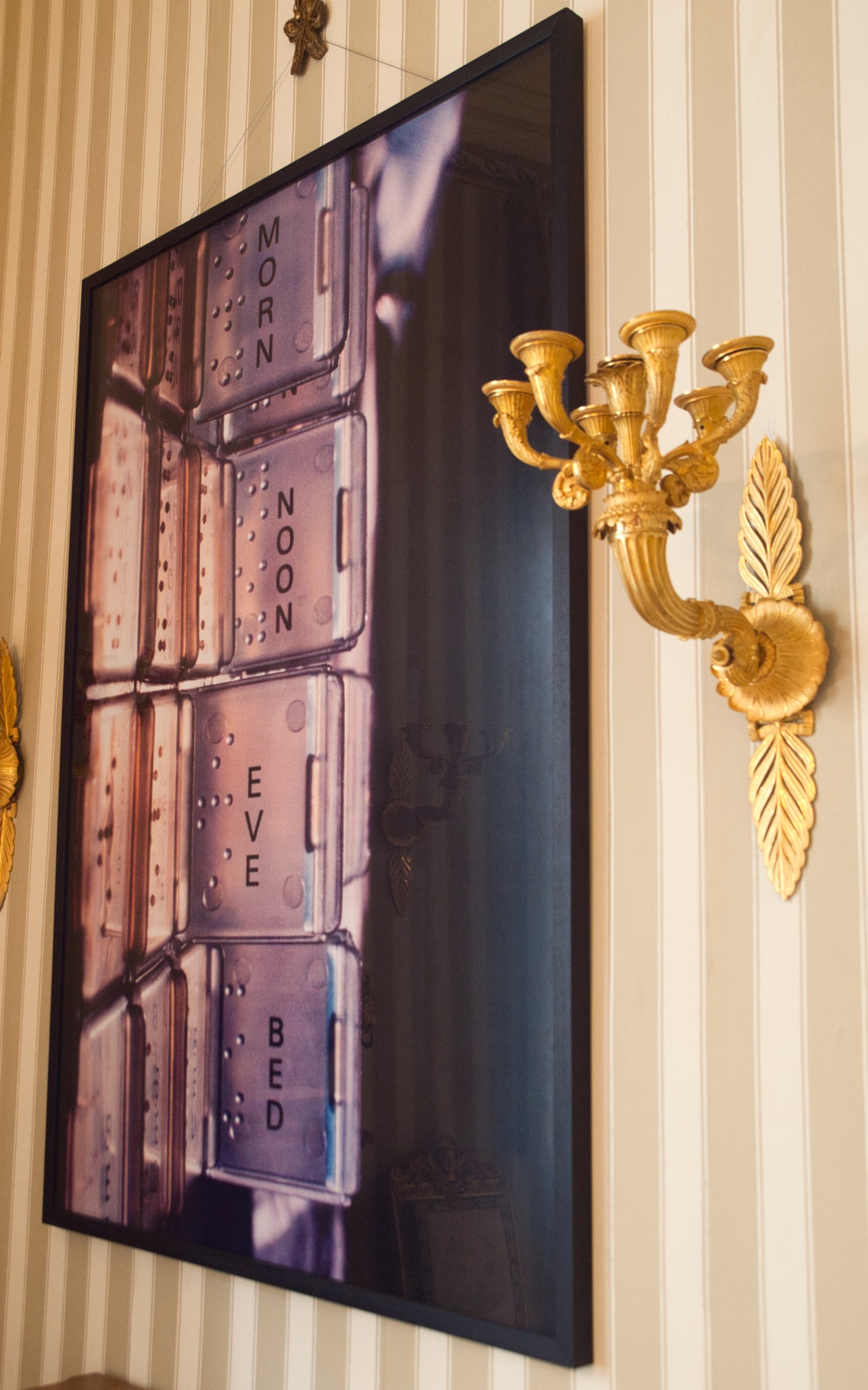L'image montre un tableau accroché au mur, représentant une série de compartiments ou de cases avec des inscriptions. Les mots "MORN," "NOON," "EVE," et "BED" sont visibles, suggérant une organisation liée à des moments de la journée. À côté du tableau, il y a un élégant applique murale en métal doré, qui ajoute une touche de sophistication à l'ensemble. Le mur est orné de rayures verticales, donnant une ambiance classique et raffinée.