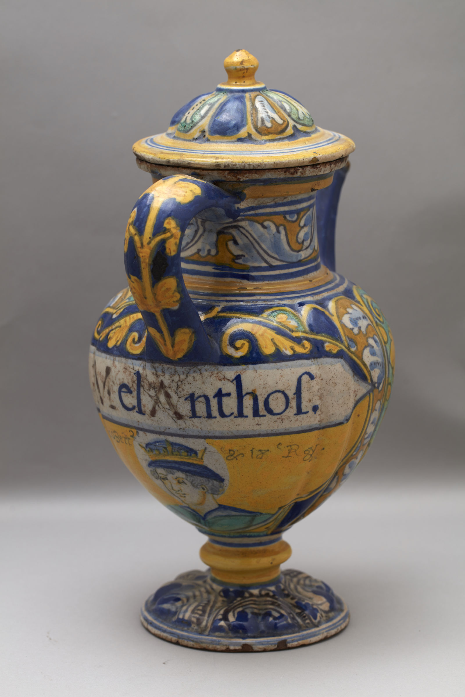 L'image montre une jarre décorative avec un couvercle, probablement en céramique. Elle présente des motifs colorés dans des tons de bleu, jaune et vert. Le corps de la jarre est richement orné de motifs floraux et de détails artistiques. On peut également voir une inscription sur le côté, suggérant un aspect historique ou culturel. La forme de la jarre, avec son pied sculpté et sa anse, ajoute à son élégance.