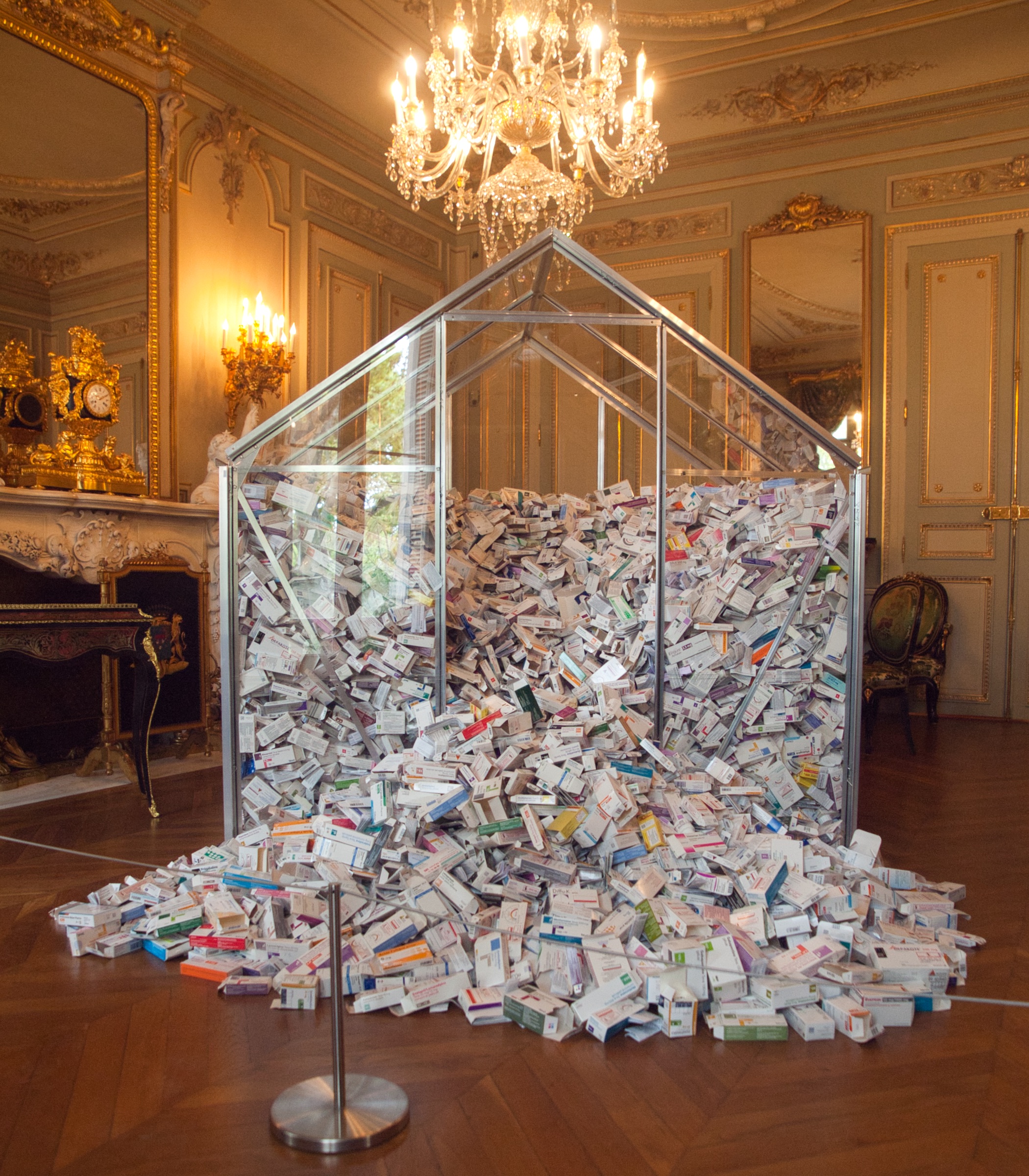 L'image montre une grande structure en forme de serre en verre, remplie de milliers de boîtes de médicaments. Cette installation est située dans une pièce élégante, ornée de miroirs et de luminaires dorés, avec un sol en parquet. Les boîtes de médicaments sont entassées à l'intérieur de la serre, créant un effet visuel impressionnant, contrastant avec le décor luxueux de la pièce.