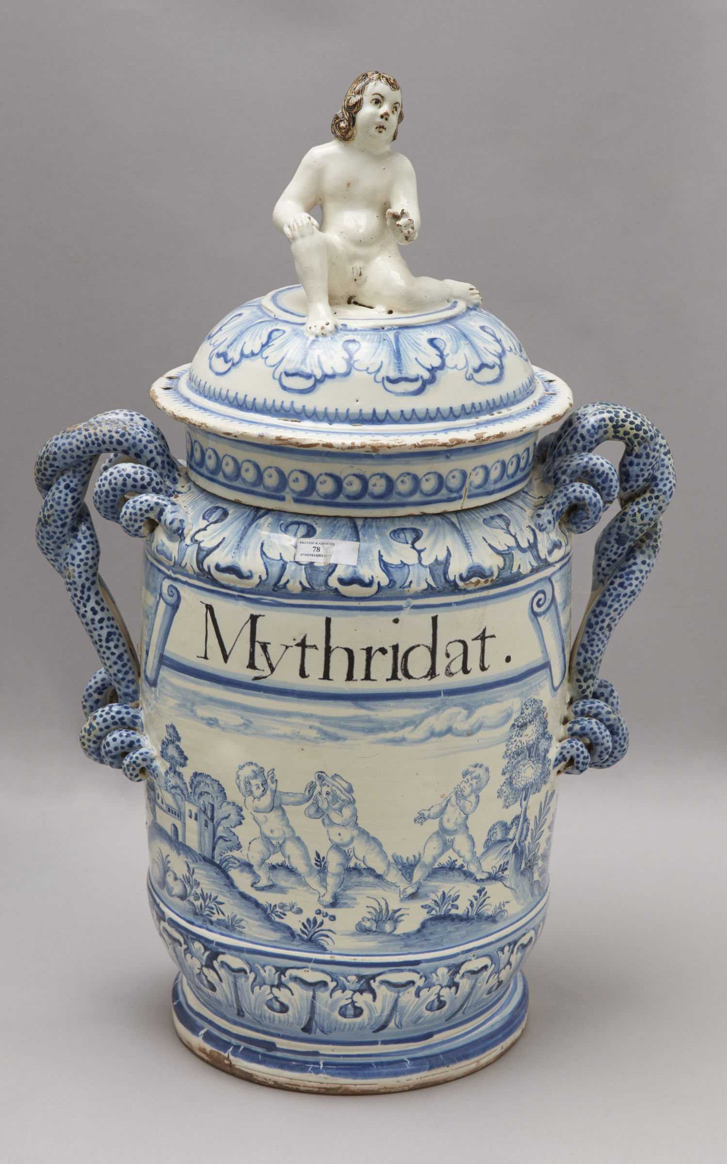 Cette image représente un grand pot ou vase en céramique, décoré de motifs bleu et blanc. Le couvercle est surmonté d'une figurine représentant un personnage nu. Sur le corps du vase, le mot "Mythridat" est inscrit, et se trouvent également des scènes peintes illustrant des motifs animaux et des paysages. Les anses latérales du vase sont ornées de motifs en relief, ajoutant à son caractère décoratif.