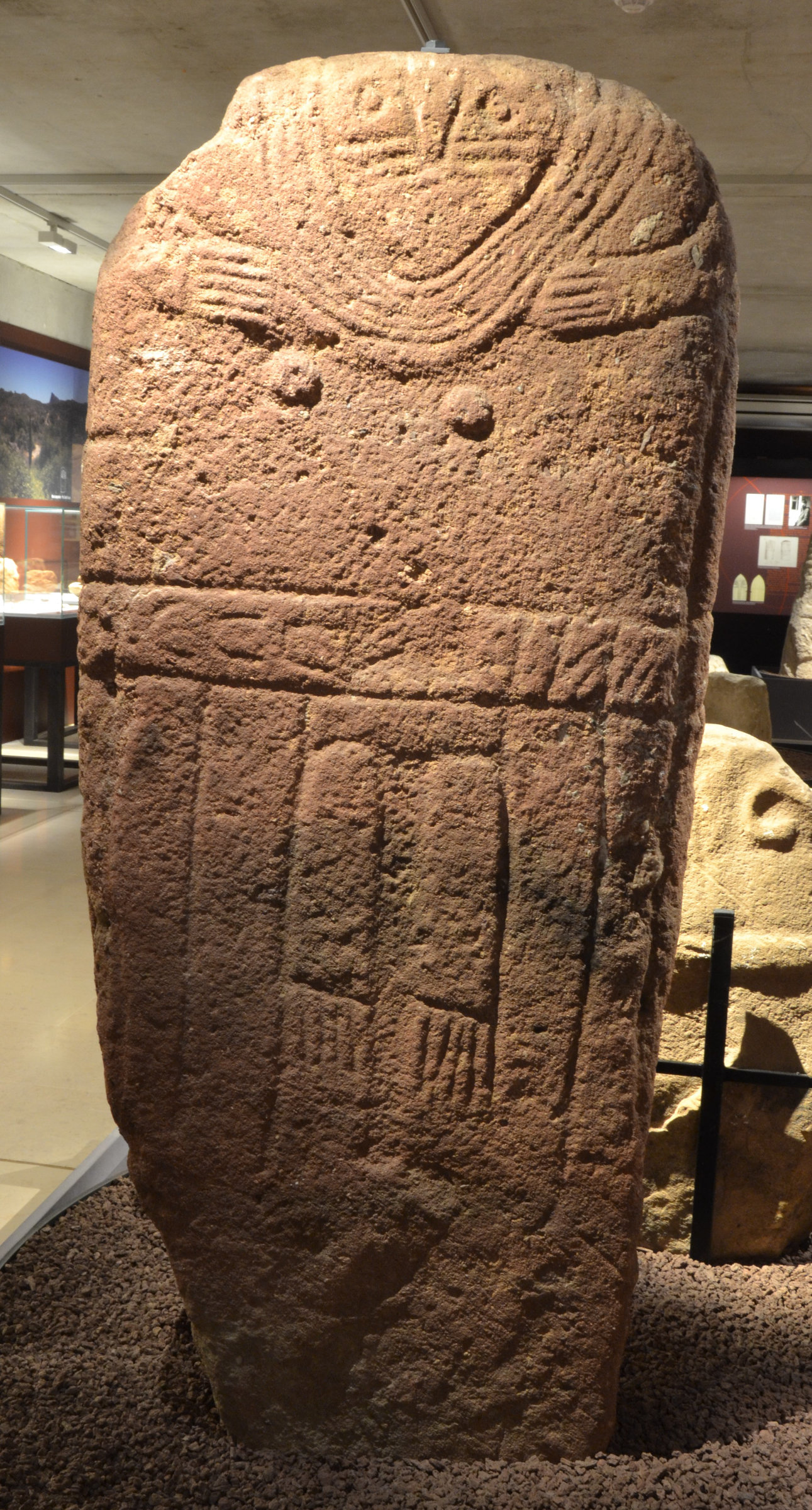 L'image montre une grande stèle en pierre, probablement ornée de motifs et de gravures. La stèle semble avoir une forme rectangulaire, avec des lignes et des figures incisées qui pourraient représenter des personnages ou des symboles symboliques. Il y a des éléments décoratifs au sommet de la pierre, ainsi que des motifs plus simples en bas. La stèle est exposée dans un cadre muséal, entourée de pierres supplémentaires, indiquant qu'il s'agit d'un artefact archéologique. Le fond montre d'autres objets exposés, suggérant un environnement de musée.
