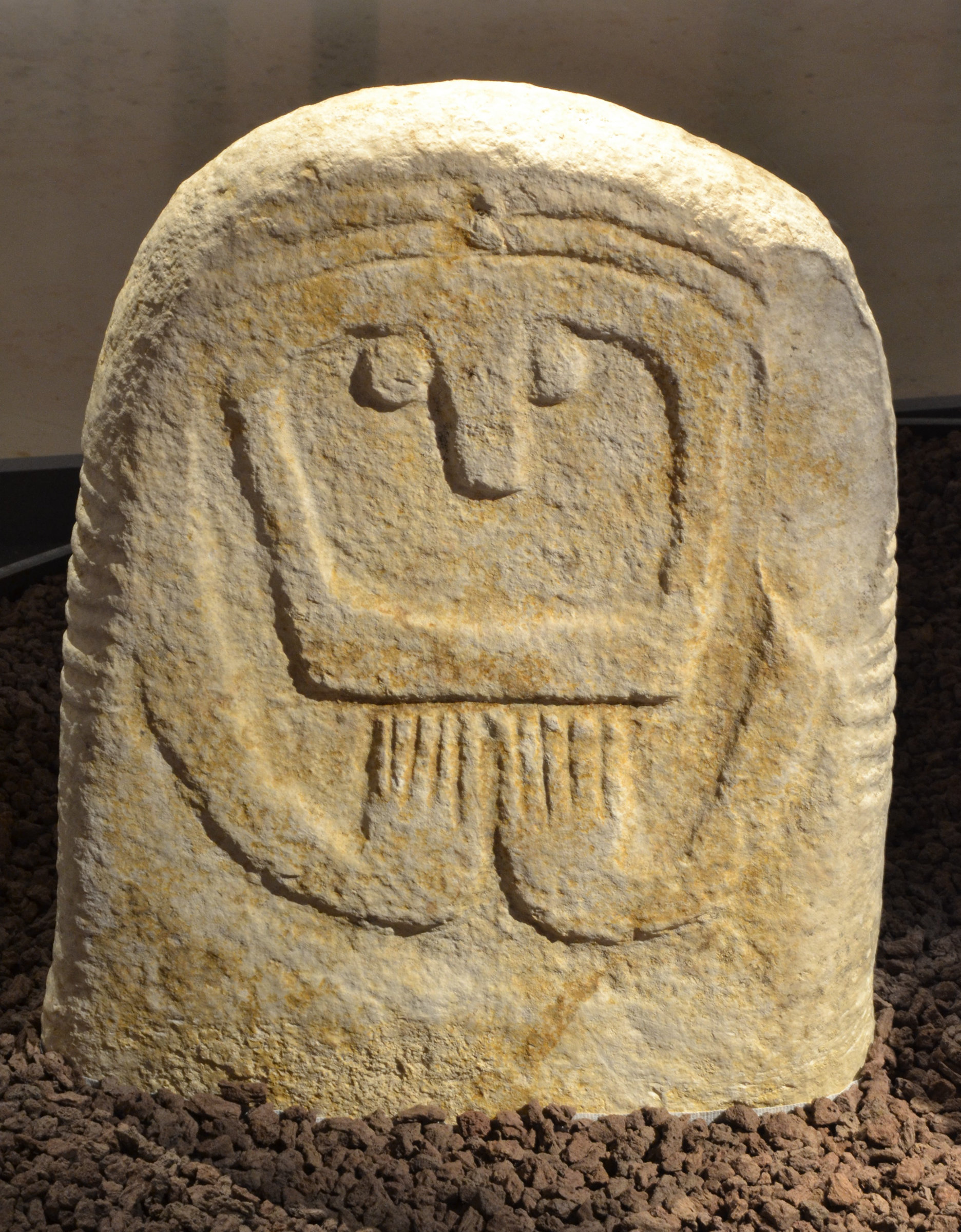 L'image montre une stèle en pierre, probablement de forme ovoïde ou rectangulaire, ornée de gravures. Ces gravures représentent une figure humaine stylisée avec un visage souriant et des bras, suggérant une posture de prière ou de vénération. La surface de la pierre est légèrement rugueuse, avec des signes d'érosion, témoignant de son ancienneté. Cette œuvre pourrait appartenir à une culture préhistorique ou ancienne, mettant en avant des éléments d'art rupestre ou de ritualisation.