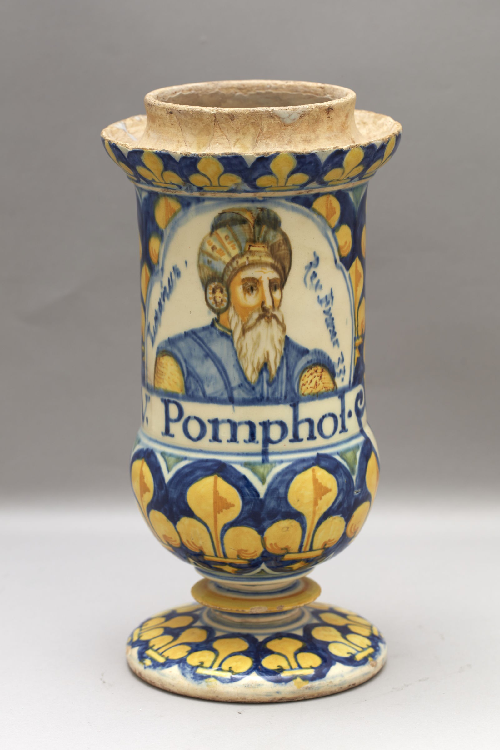 Cette image représente un vase décoratif en céramique. Il est orné de motifs floraux allant du bleu au jaune. Au centre, il y a un portrait d'un homme avec une barbe et un turban, accompagné de l'inscription "Pomphoi". Le vase a une forme élargie vers le haut et repose sur un pied. Les détails de la peinture ajoutent une touche d'élégance à l'ensemble de l'objet.