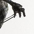 L'image semble représenter une partie d'une sculpture, mettant en avant une main réaliste et détaillée. La main est légèrement courbée et tendue, avec des doigts qui semblent interagir avec un fil ou une corde. La texture de la sculpture suggère un travail minutieux, probablement en métal ou en bronze, avec des ombres qui ajoutent de la profondeur. Le fond est clair, ce qui permet de mettre en valeur la forme et les détails de la main.
