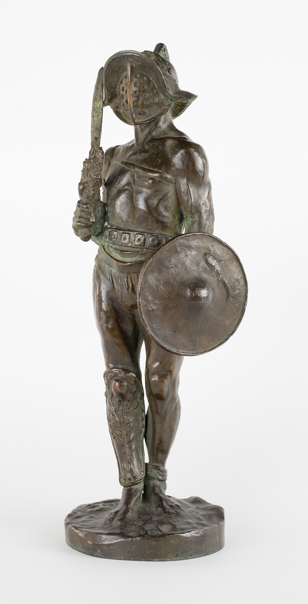 L'image représente une sculpture en bronze d'un guerrier. Il est sculpté en position debout, tenant un glaive dans une main et un bouclier dans l'autre. Le guerrier est musclé, avec des détails anatomiques très marqués. Il porte un casque et des éléments d'armure sur ses jambes, tandis que son corps est nu sur la majorité. La sculpture est sur un socle, ce qui lui confère une certaine prestance. L'ensemble de l'œuvre dégage une impression de force et de bravoure.