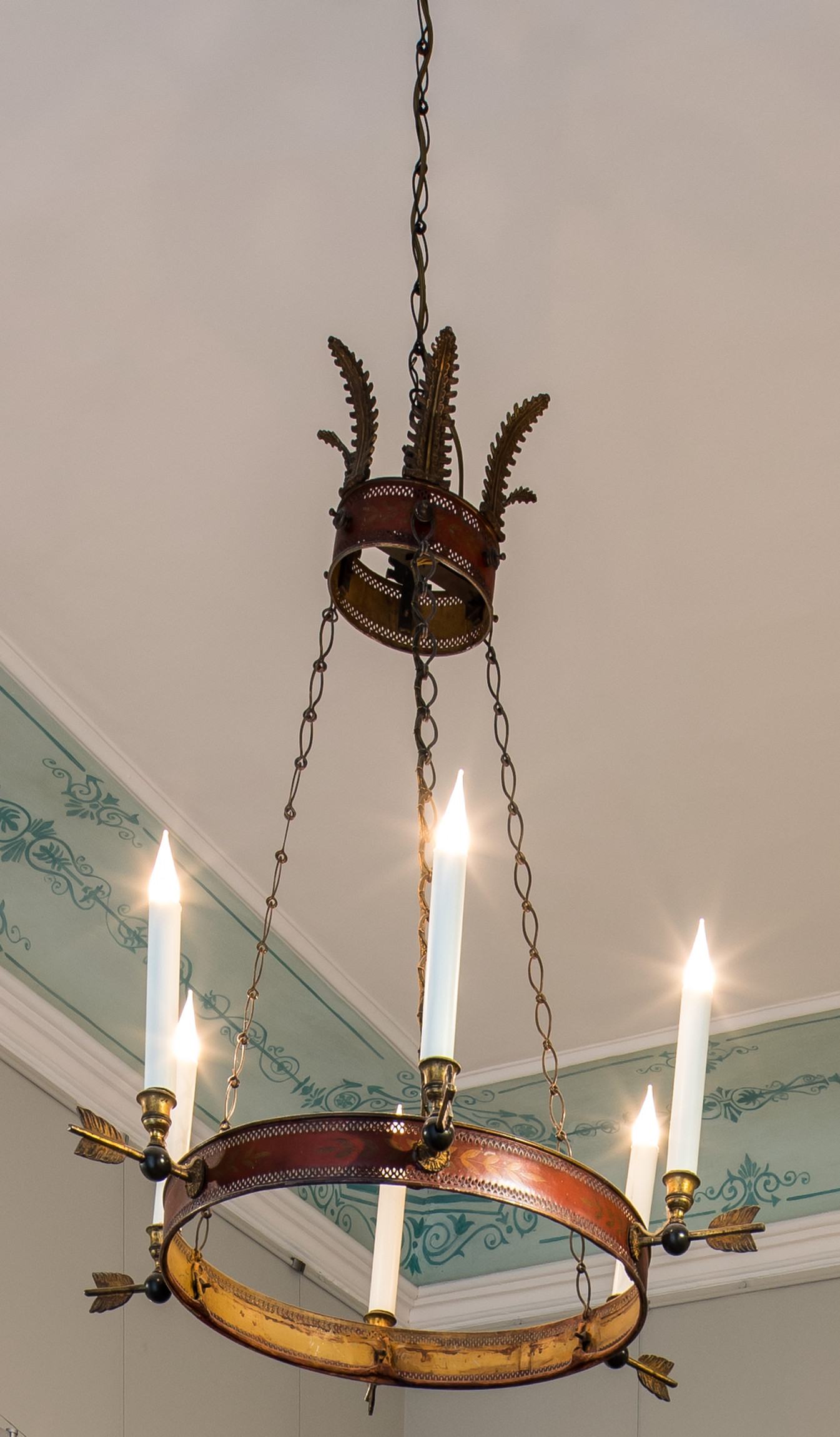 L'image montre un lustre élégant suspendu au plafond. Il est composé d'une structure en métal, probablement en fer forgé, et comporte plusieurs bras qui accueillent des bougies. Les bougies sont de forme allongée et de couleur blanche. Le design du lustre choisit des motifs ornementaux, avec des détails décoratifs sur la base. Le plafond, visible en arrière-plan, présente une décoration peinte dans des teintes douces qui complètent l'esthétique du lustre.